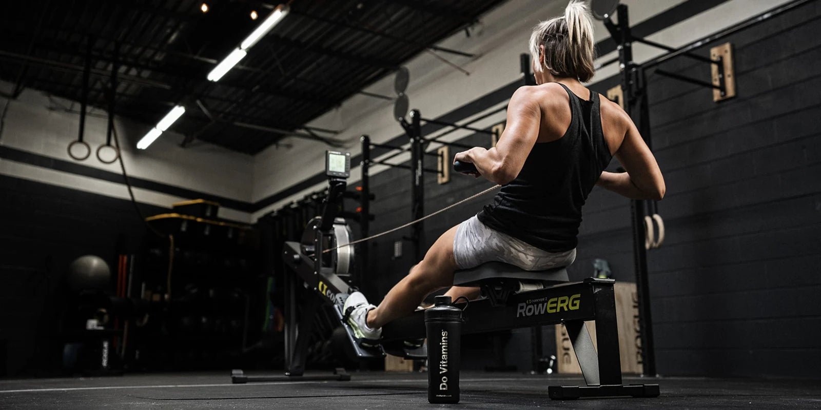 An athlete rowing in a gym, with a Do Vitamins shaker beside her